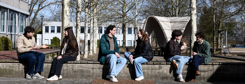 Studierende sitzen auf einer Sitzfläche am Campus und unterhalten sich.