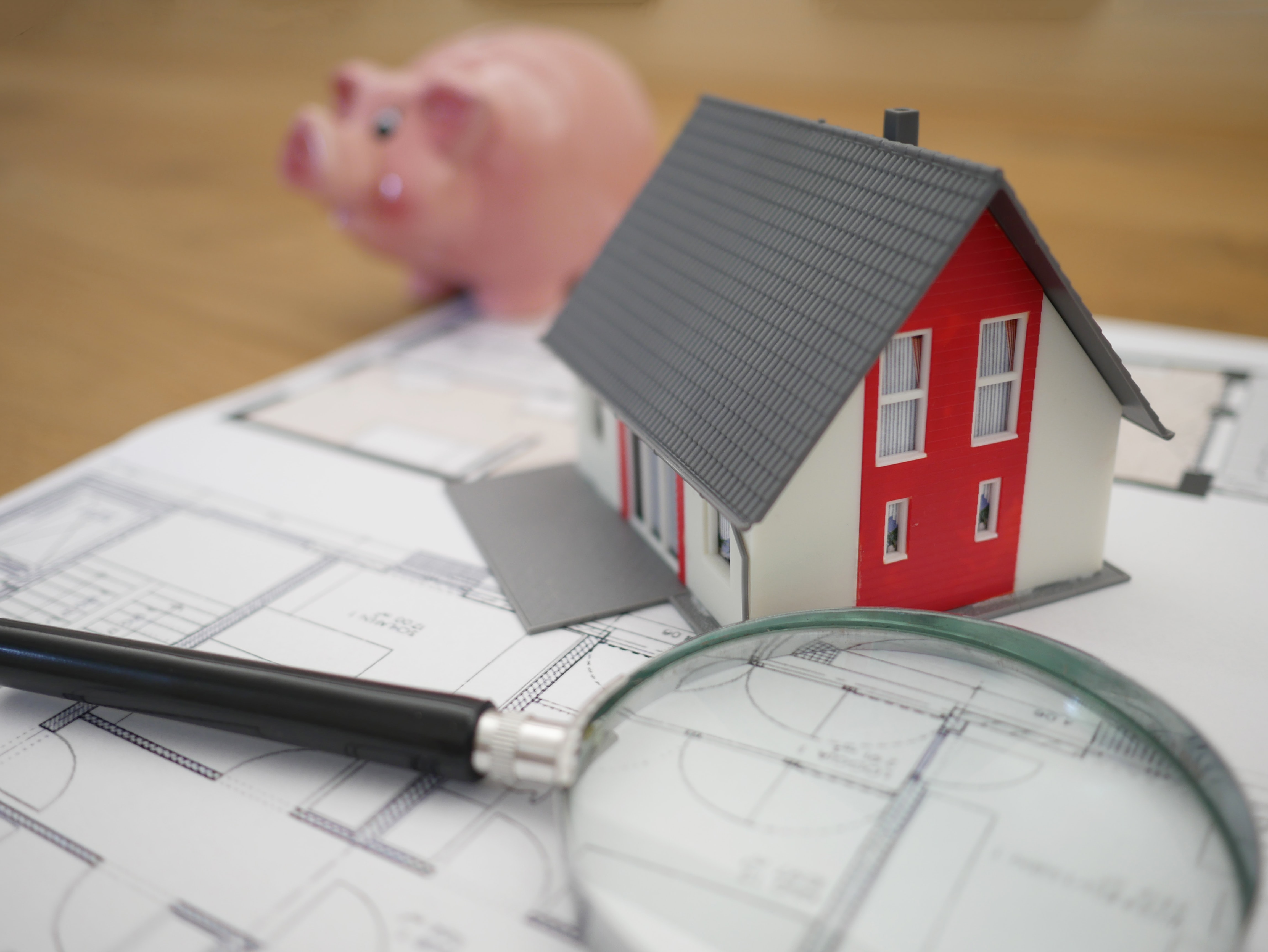 Wohnungsmarkt (Modul 24) Ein Modellhaus steht auf einem Bauplan mit einer Lupe und einem Sparschwein daneben.