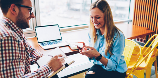 Besprechungssituation Ein Mann mit Notizblock und Stift und eine Frau mit Handy sitzen sich gegenüber und lächeln entspannt. An ihrer Seite steht ein Tisch mit geöffnetem Laptop. Die Stühle sind gelb.