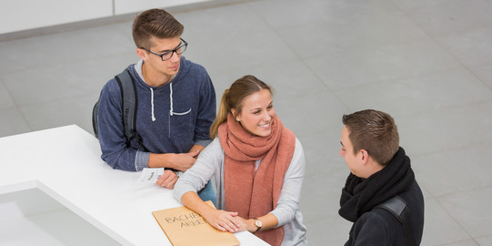 Studierendensekretariat Drei Personen stehen neben einenander an einer Theke. Die Person in der Mitte lacht die Person rechts an und hat ihre Hände auf einen großen Umschlag liegen. Die Person links hat einen kleinen Zettel mit einer Wartenummer in der Hand.